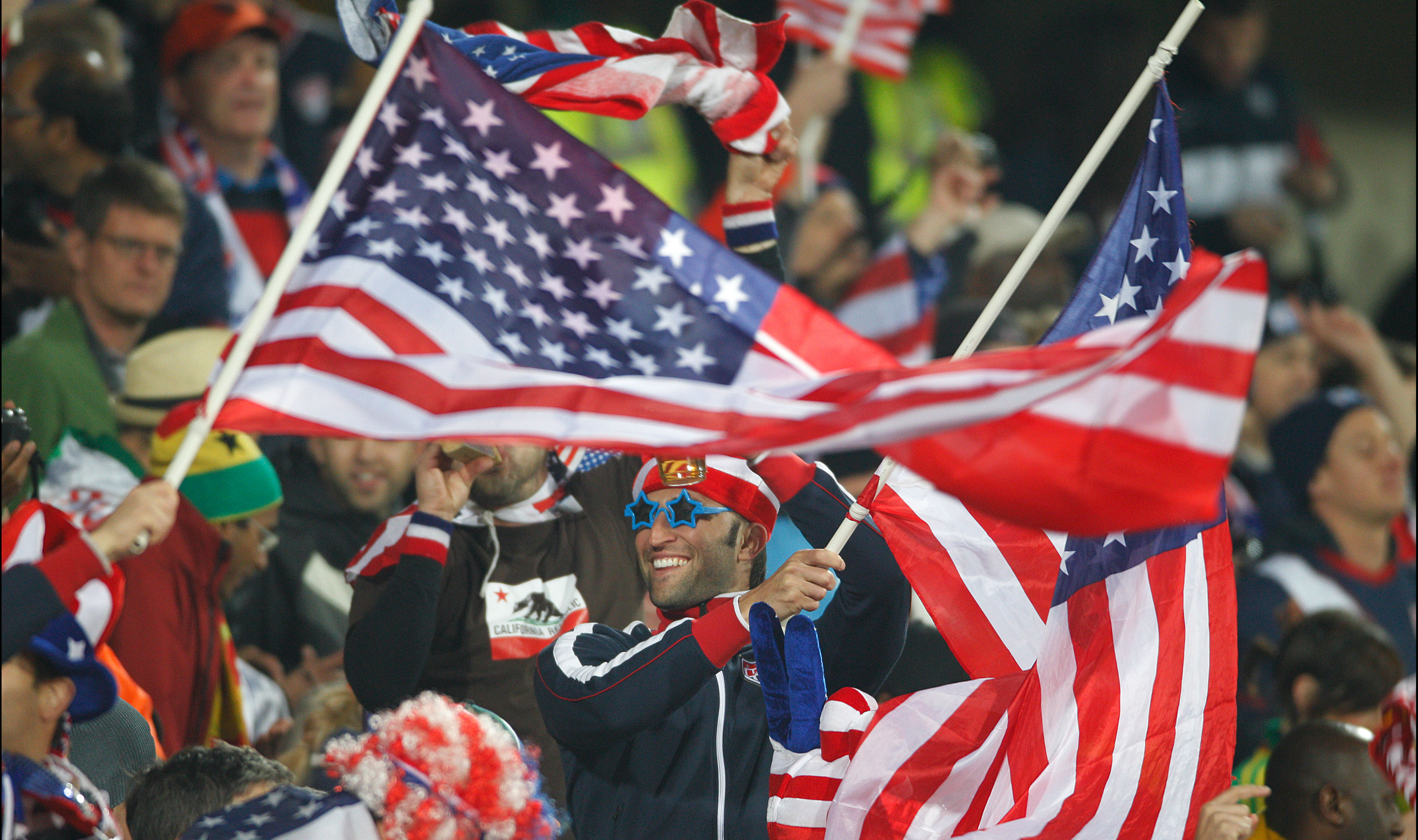 Smiling fan in blue sunglasses waves two American flags among a crowd of supporters.