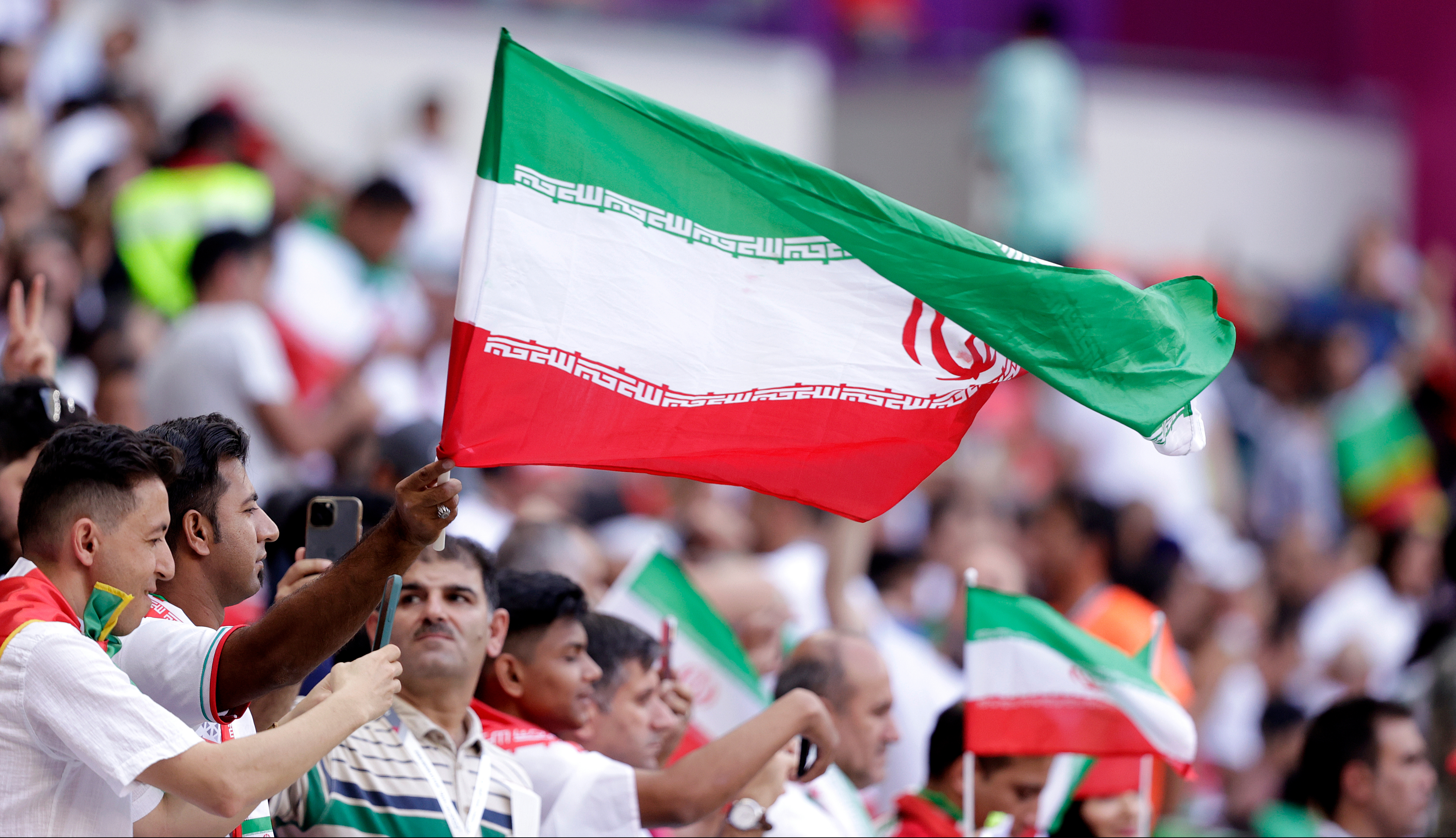 A vibrant Iranian flag with green, white, and red stripes waves above a cheering crowd.