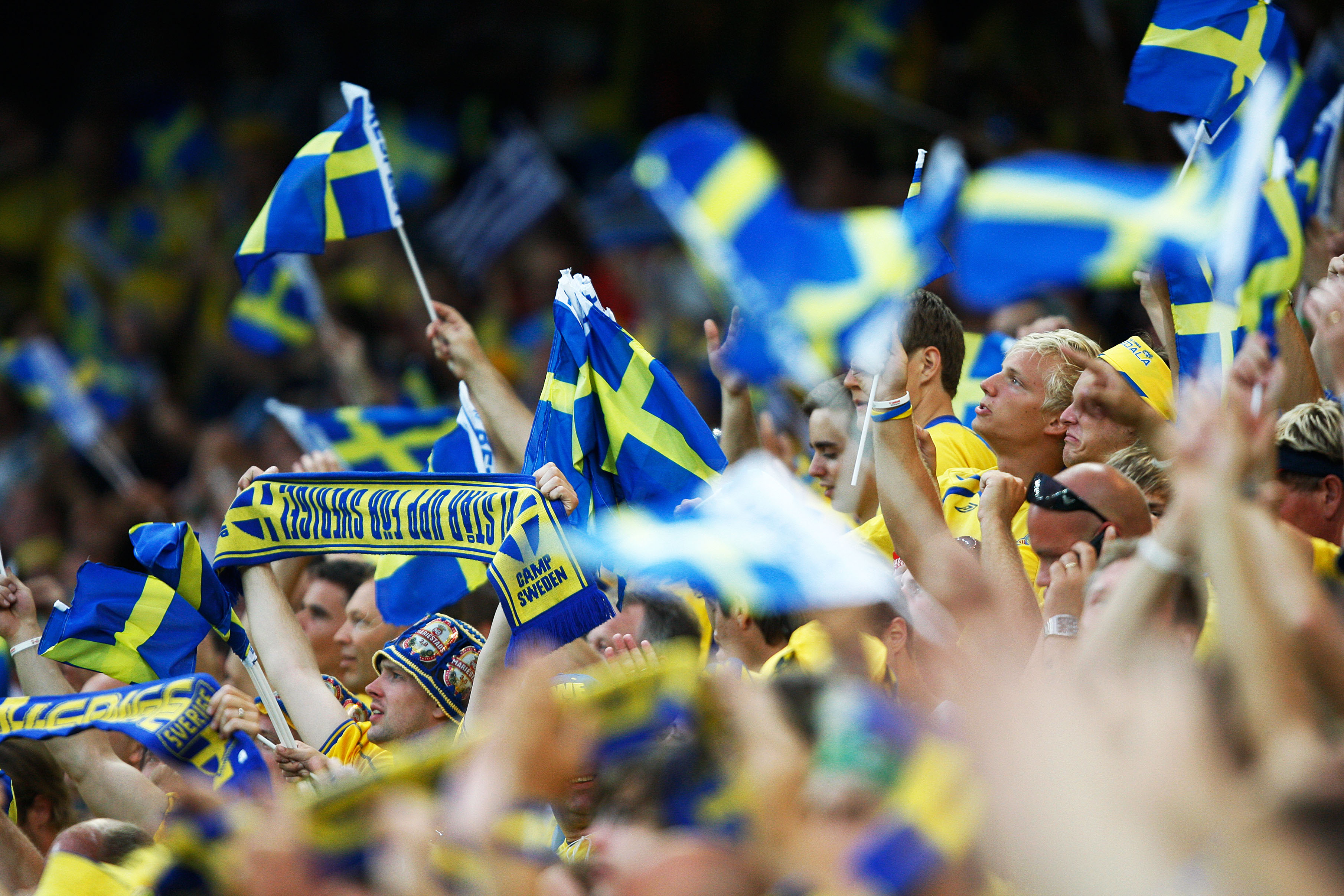 A crowd of enthusiastic fans wave numerous blue and yellow Swedish flags at an event.