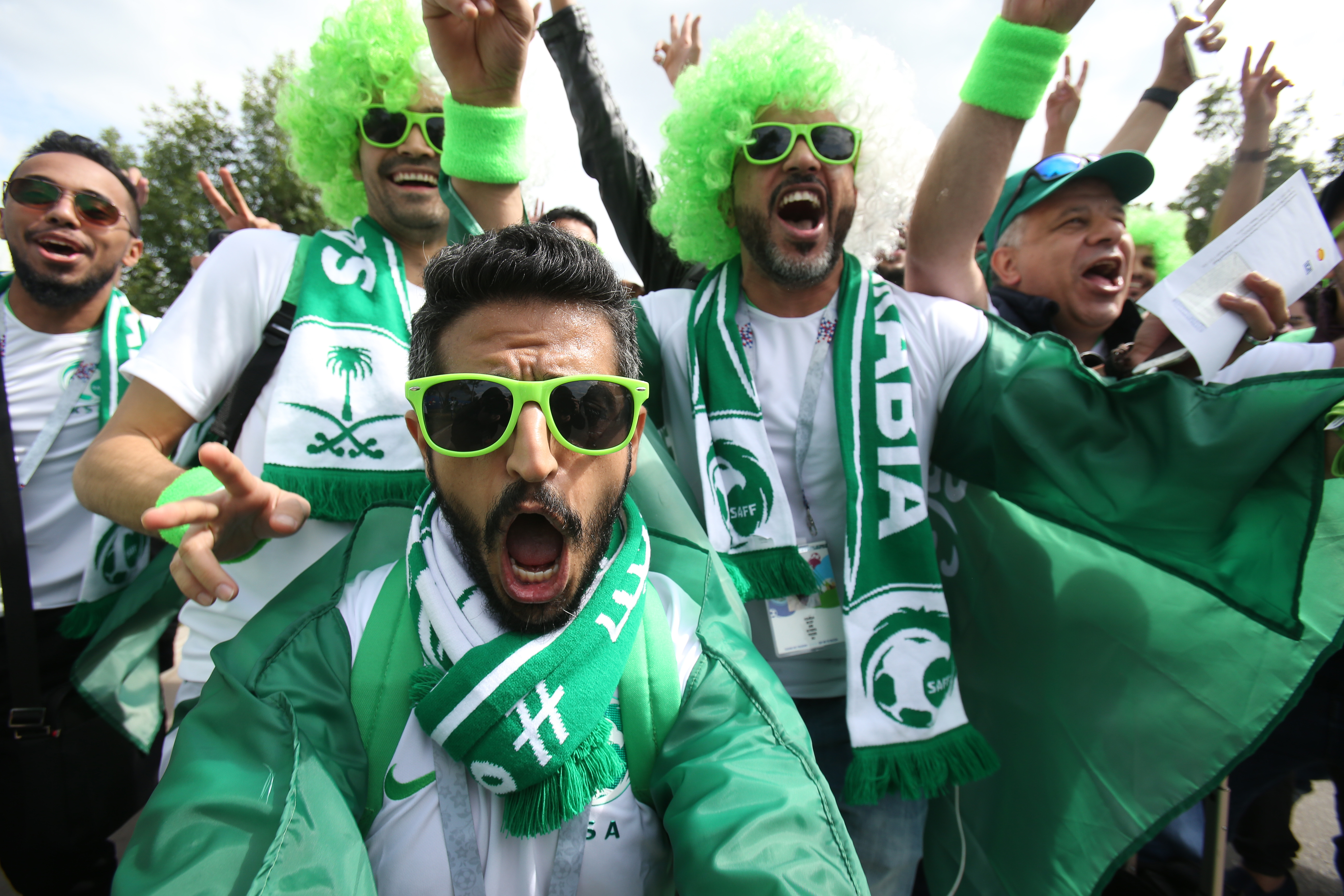 Excited Saudi football fans in green wigs and scarves cheer loudly at an event.