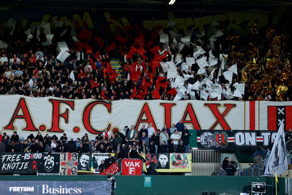 Passionate football fans in a stadium display a large banner reading "A.F.C. AJAX".