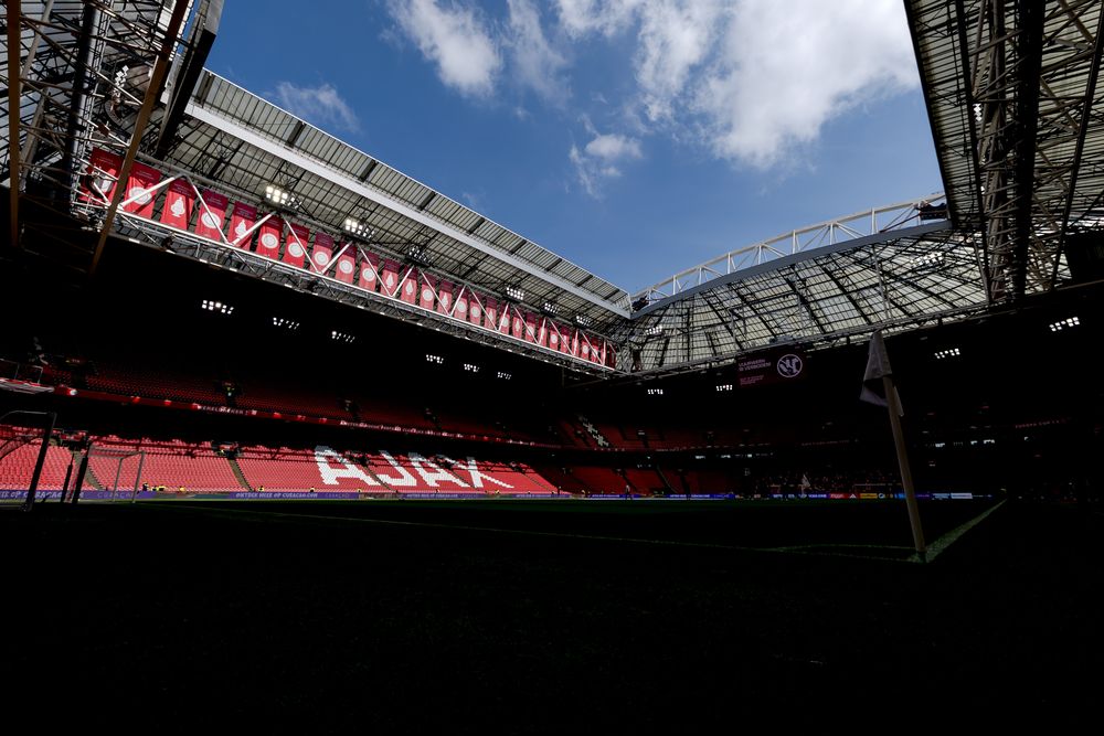 Empty stadium interior with red 'AJAX' seating under a bright blue sky.