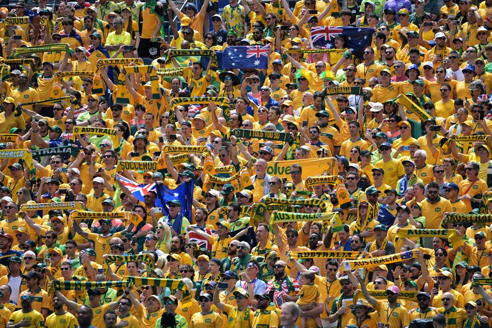 Large crowd of Australian fans in yellow shirts cheering and waving flags enthusiastically.