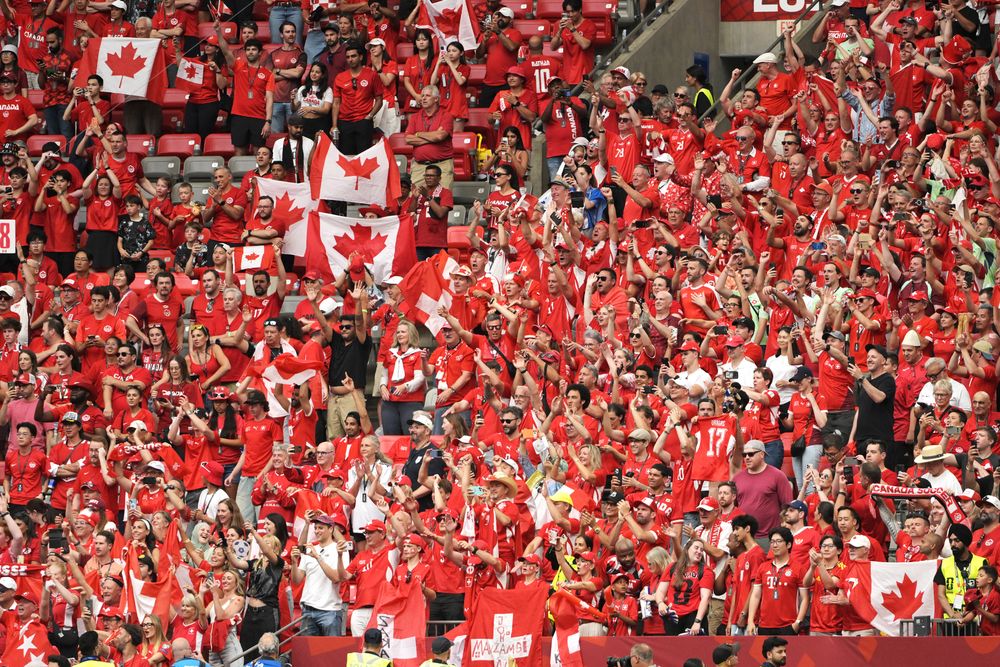 A large stadium crowd, mostly dressed in red, waving Canadian flags and cheering.