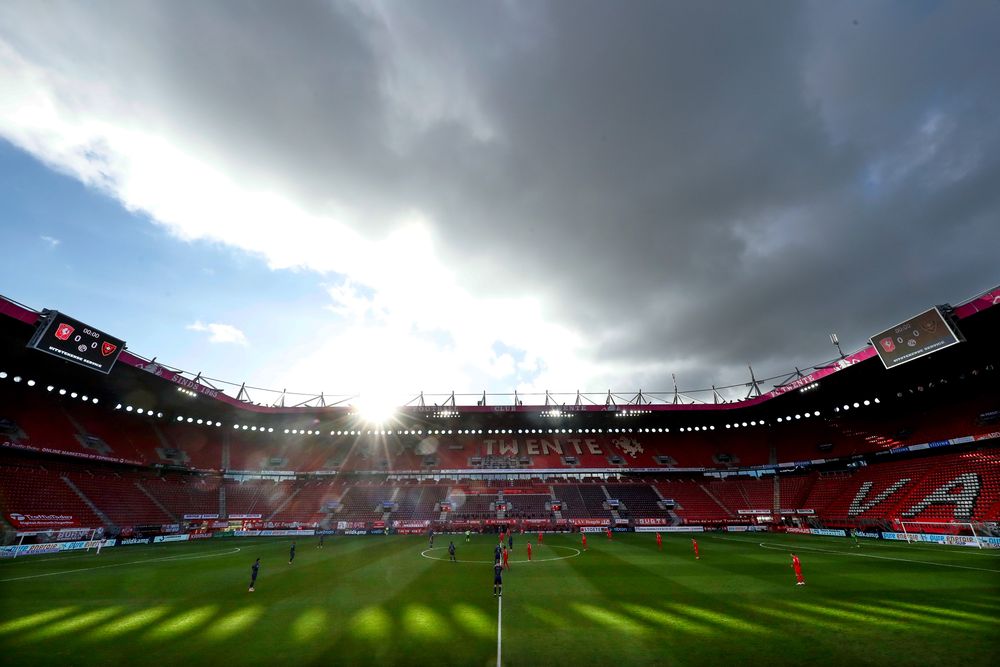 Football stadium with a dramatic sky, bright sunlight, green pitch, and red seating.