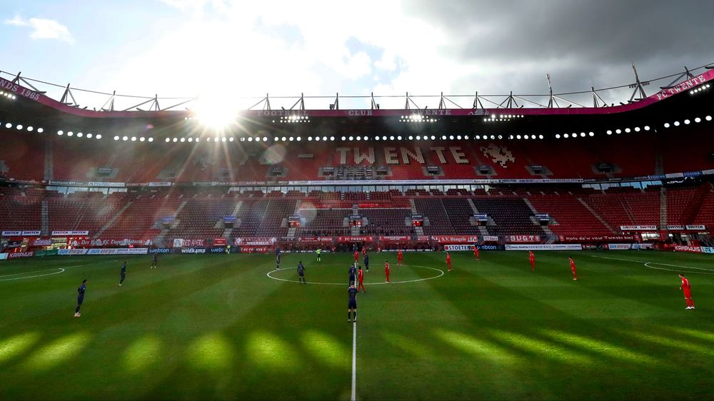Football stadium with a dramatic sky, bright sunlight, green pitch, and red seating.