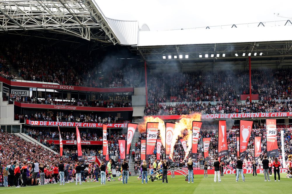 A crowded soccer stadium with fans cheering as pyrotechnics ignite on the field.