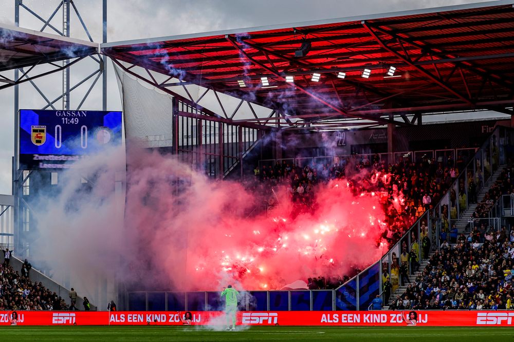 Red flares and smoke engulf a section of a football stadium with fans cheering.