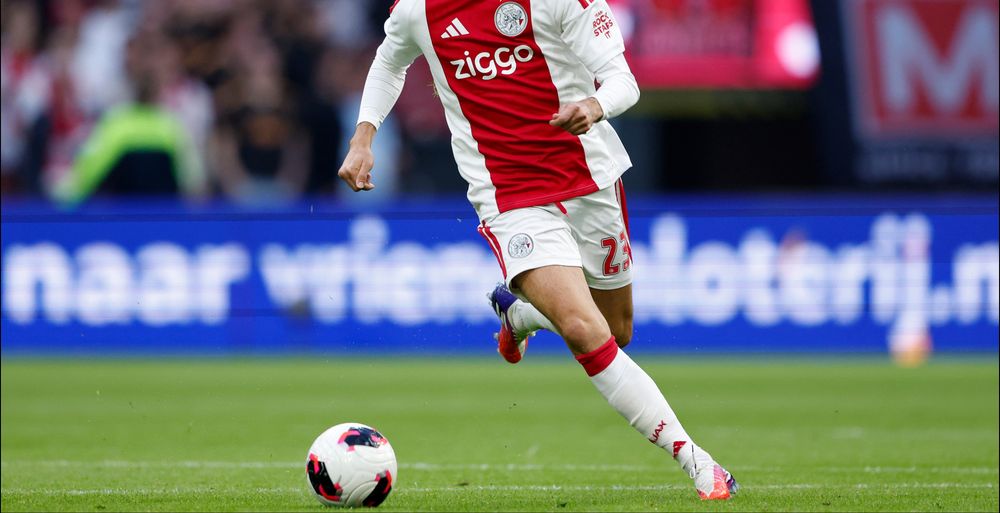 Soccer player in a red and white Ajax jersey dribbling a ball on a green field.