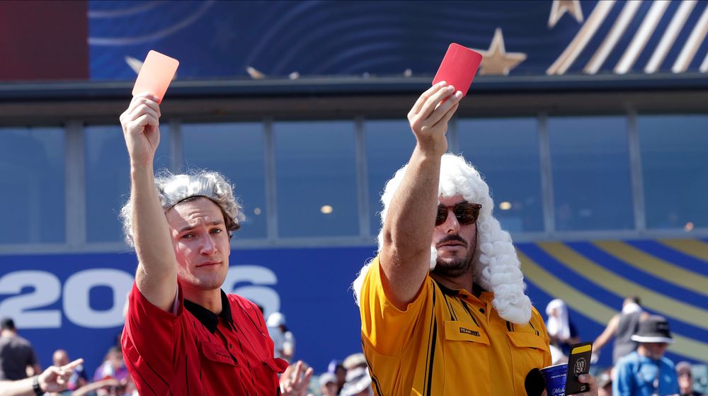 Two men in referee outfits hold red cards up against a stadium background.