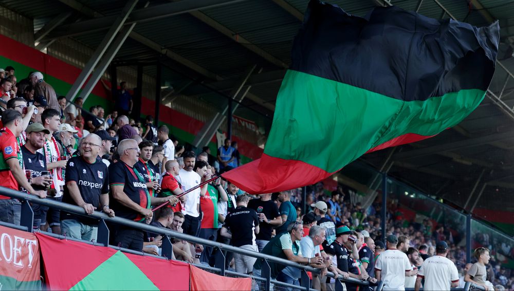 Spectators in a stadium watch a game, a large black, green, and red flag waves.