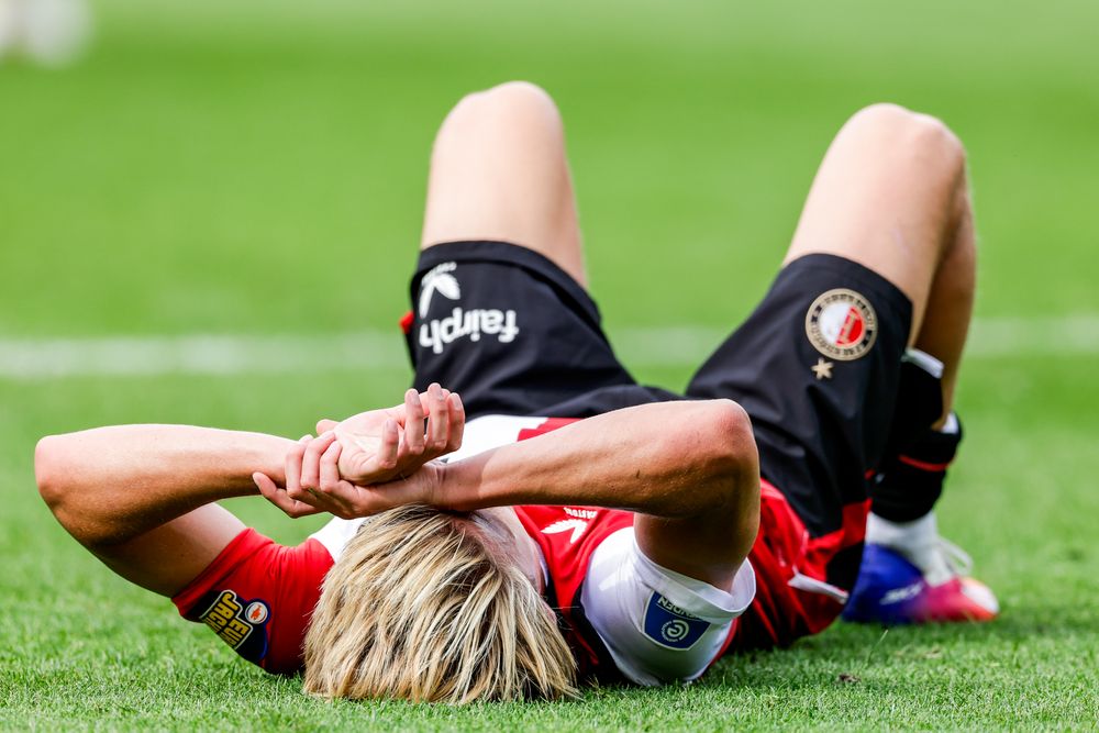 A blonde soccer player lies on the grass, hands behind head, looking defeated.