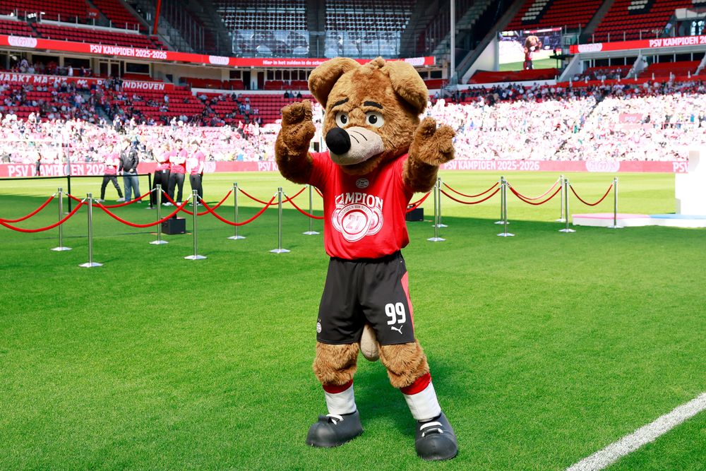 Brown bear mascot in red shirt and black shorts dancing on a green soccer field.