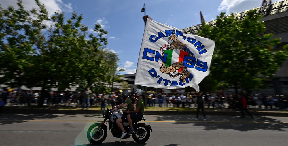 Motorcyclist waves a large white flag with 'CAMPIONI D'ITALIA' text under a sunny sky.