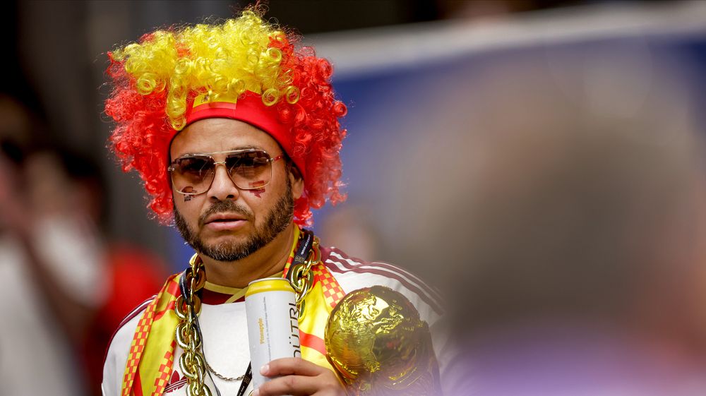 Man with red and yellow wig, sunglasses, gold chain, holding golden trophy.