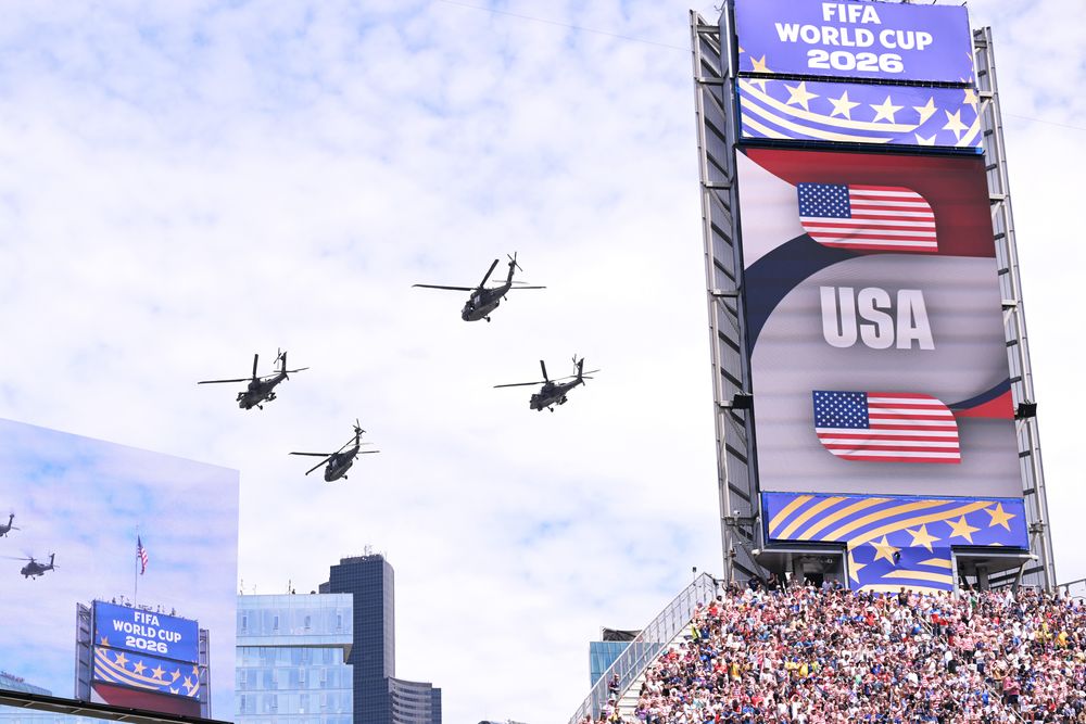 Several helicopters fly past a stadium jumbotron displaying 'FIFA WORLD CUP 2026' and US flags.