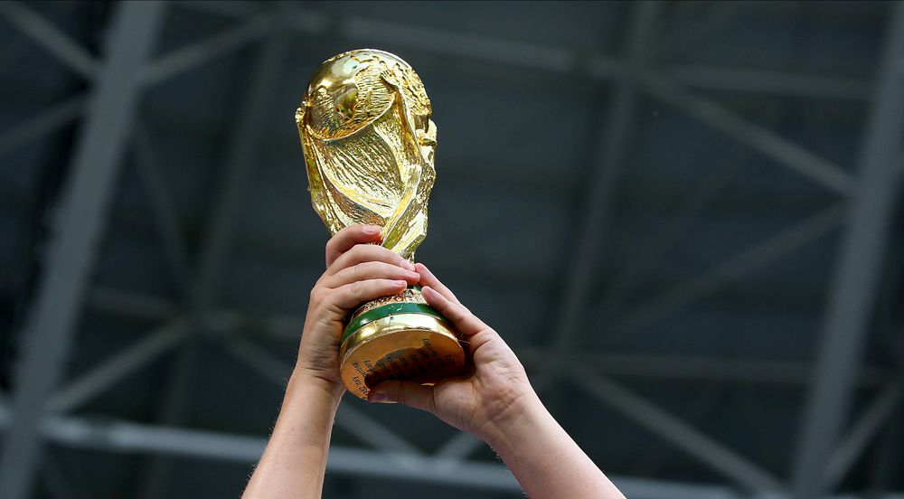 Two hands hold up the golden FIFA World Cup trophy against a blurred background.