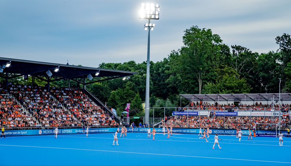 Field hockey game on blue turf, players, and spectators under stadium lights.
