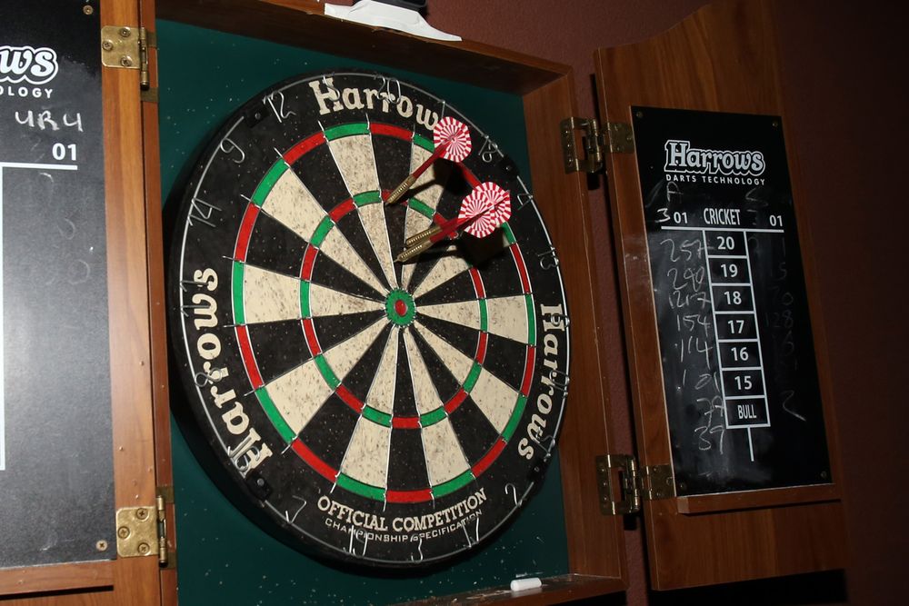 Harrows dartboard with two pink darts stuck near the center, mounted in a wooden cabinet.