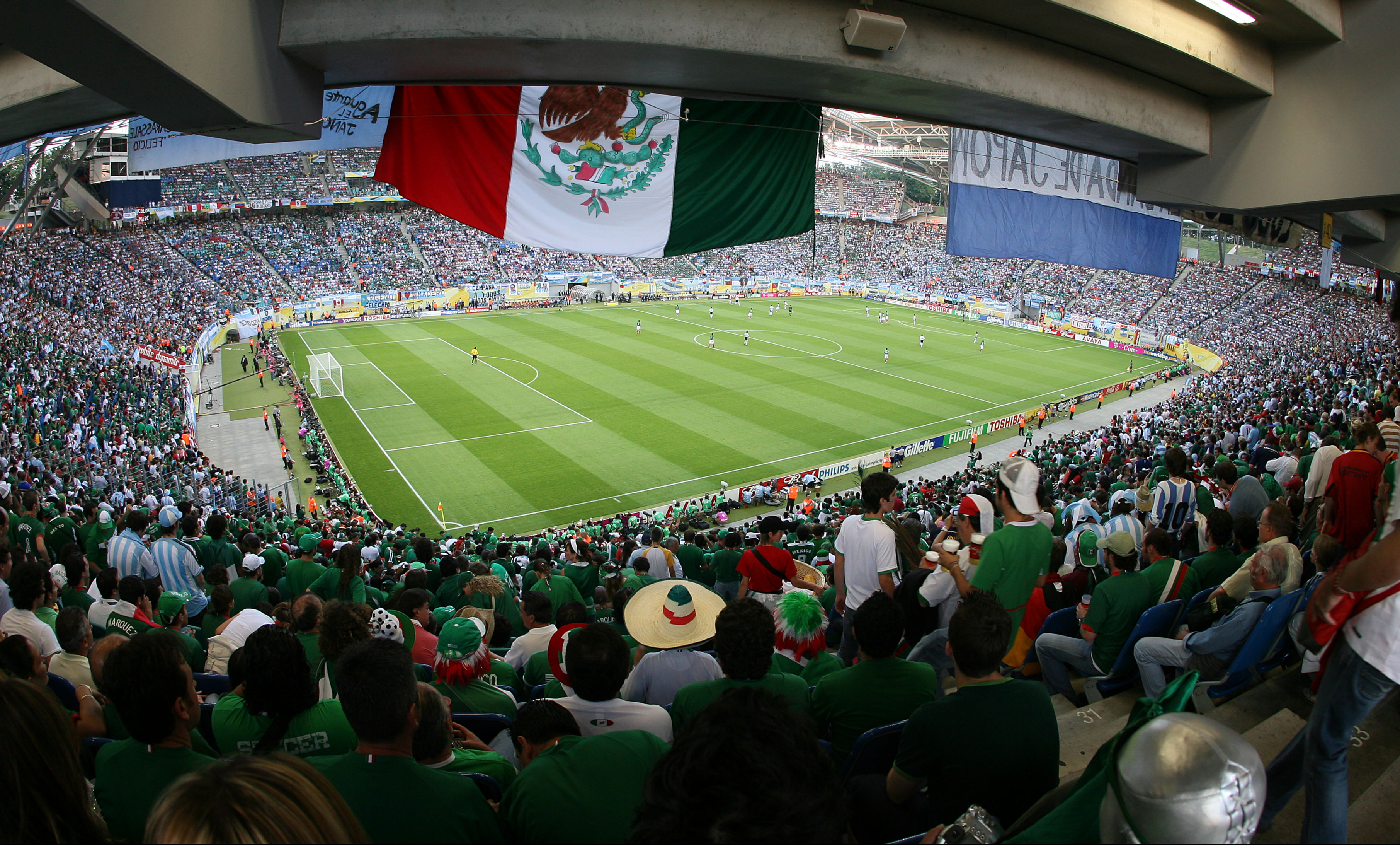 Packed soccer stadium with a large Mexican flag hanging above the enthusiastic crowd.