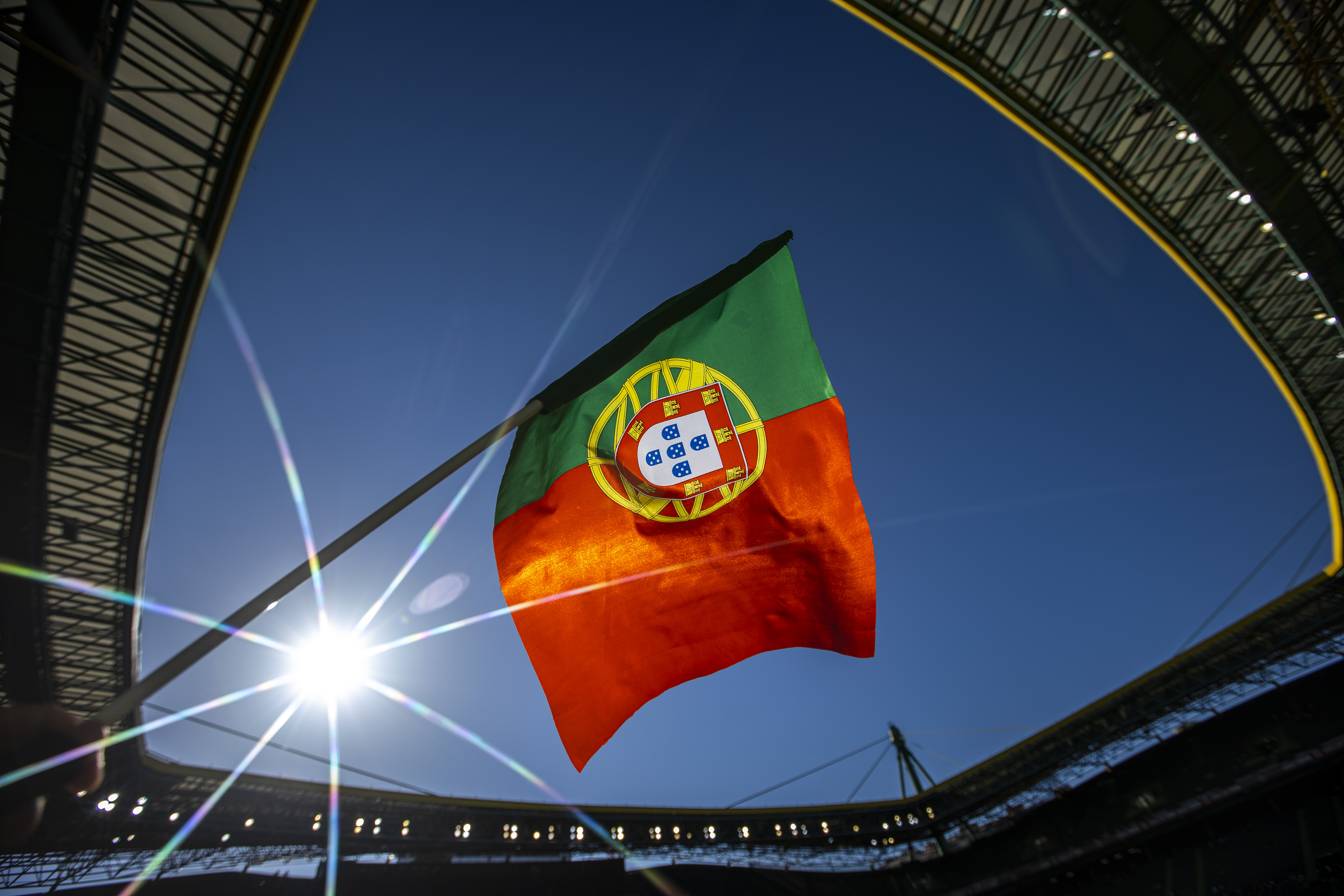 Portuguese flag waves brightly in a stadium against a clear blue sky with sun flare.