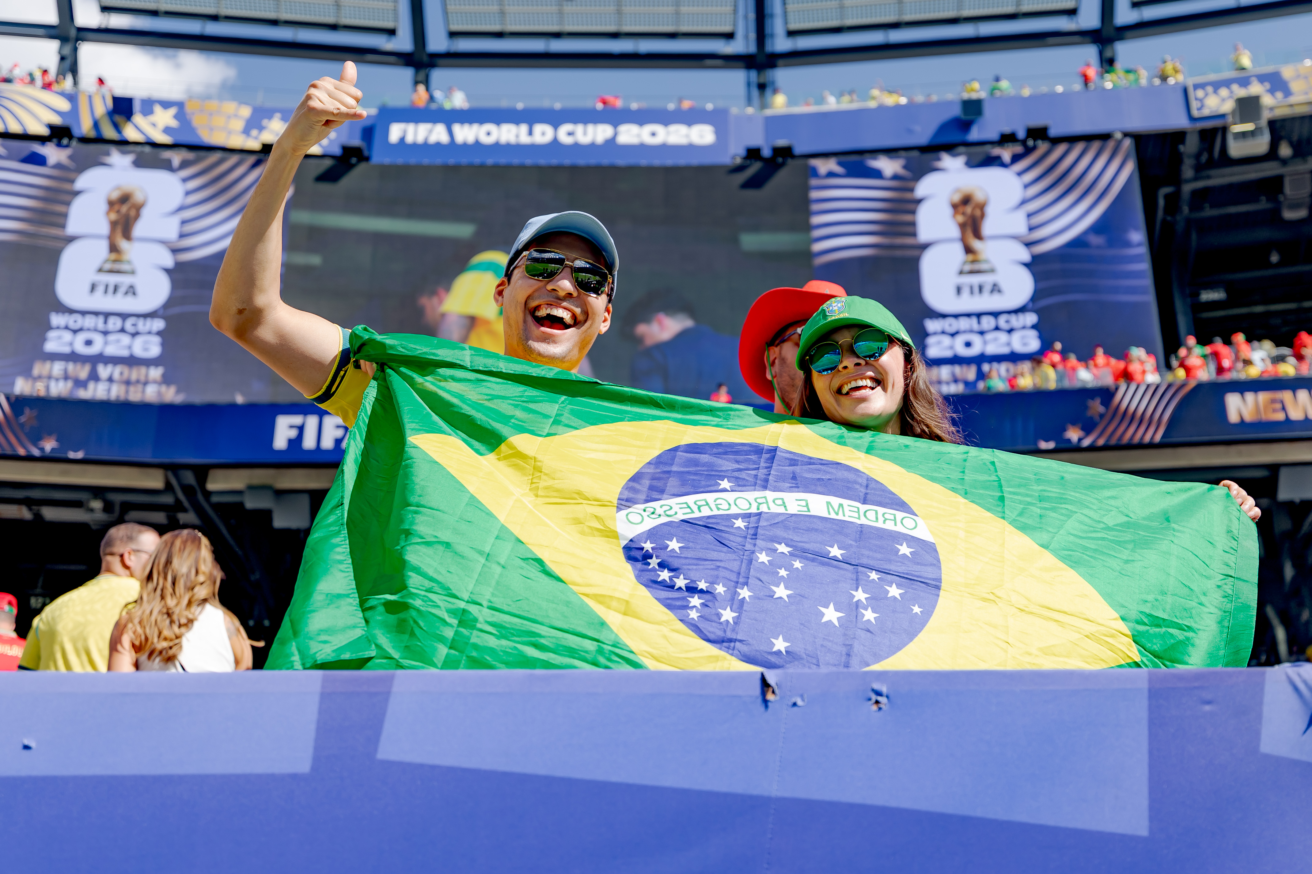 Two smiling fans proudly hold a Brazilian flag at a FIFA World Cup stadium event.