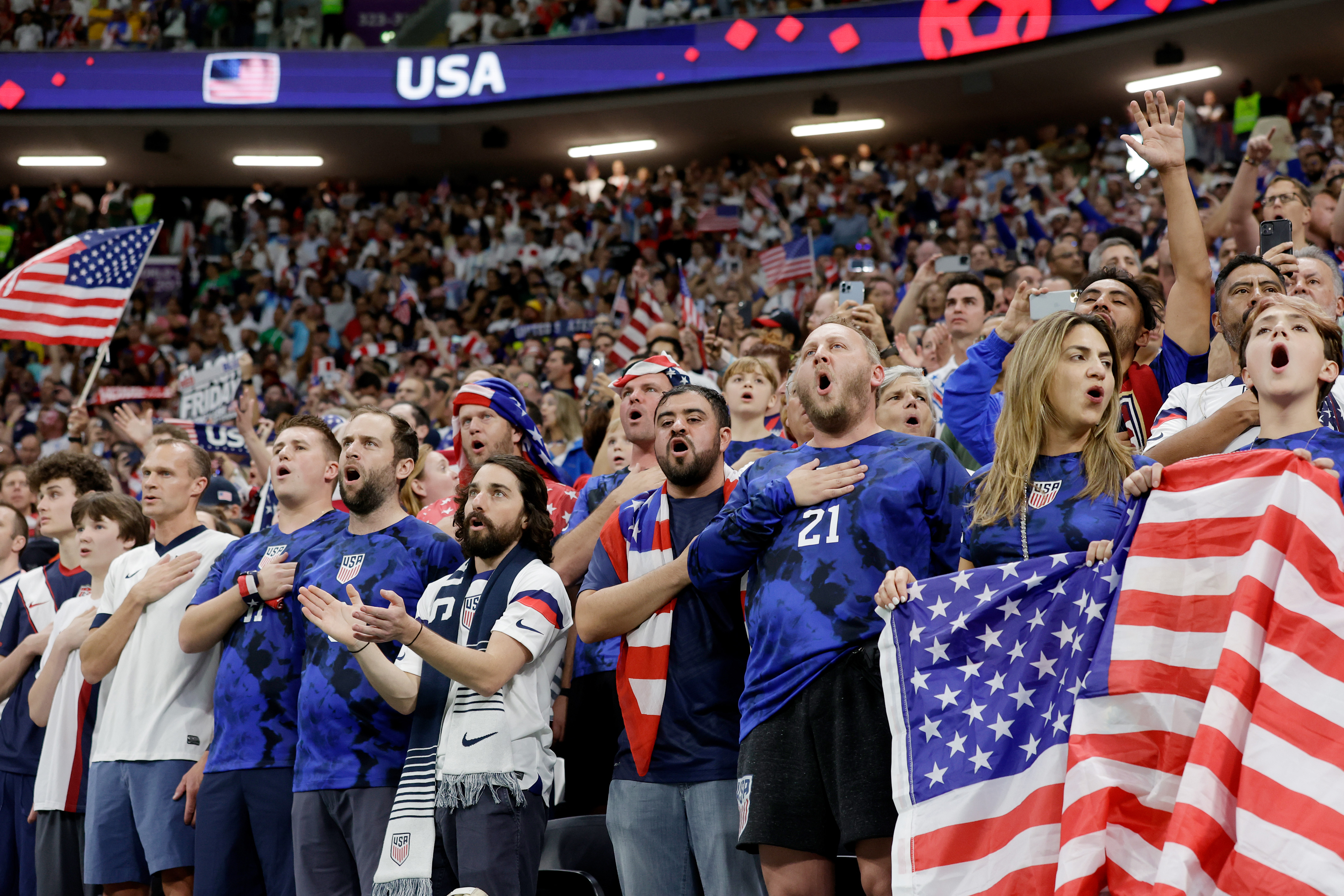 USA soccer fans passionately cheer in a stadium, many holding American flags.