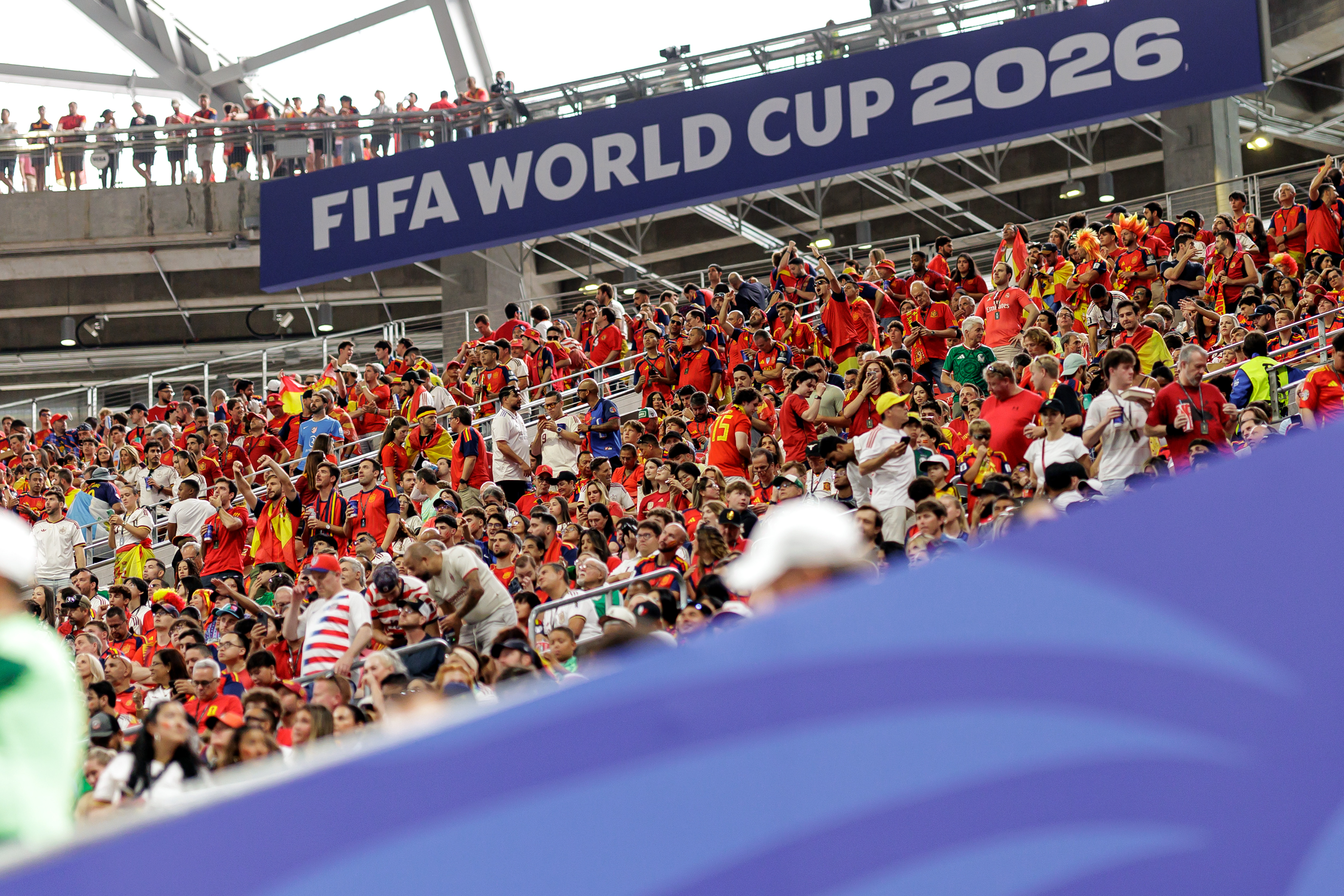 Excited soccer fans wearing red and yellow cheer under a 'FIFA World Cup 2026' banner.