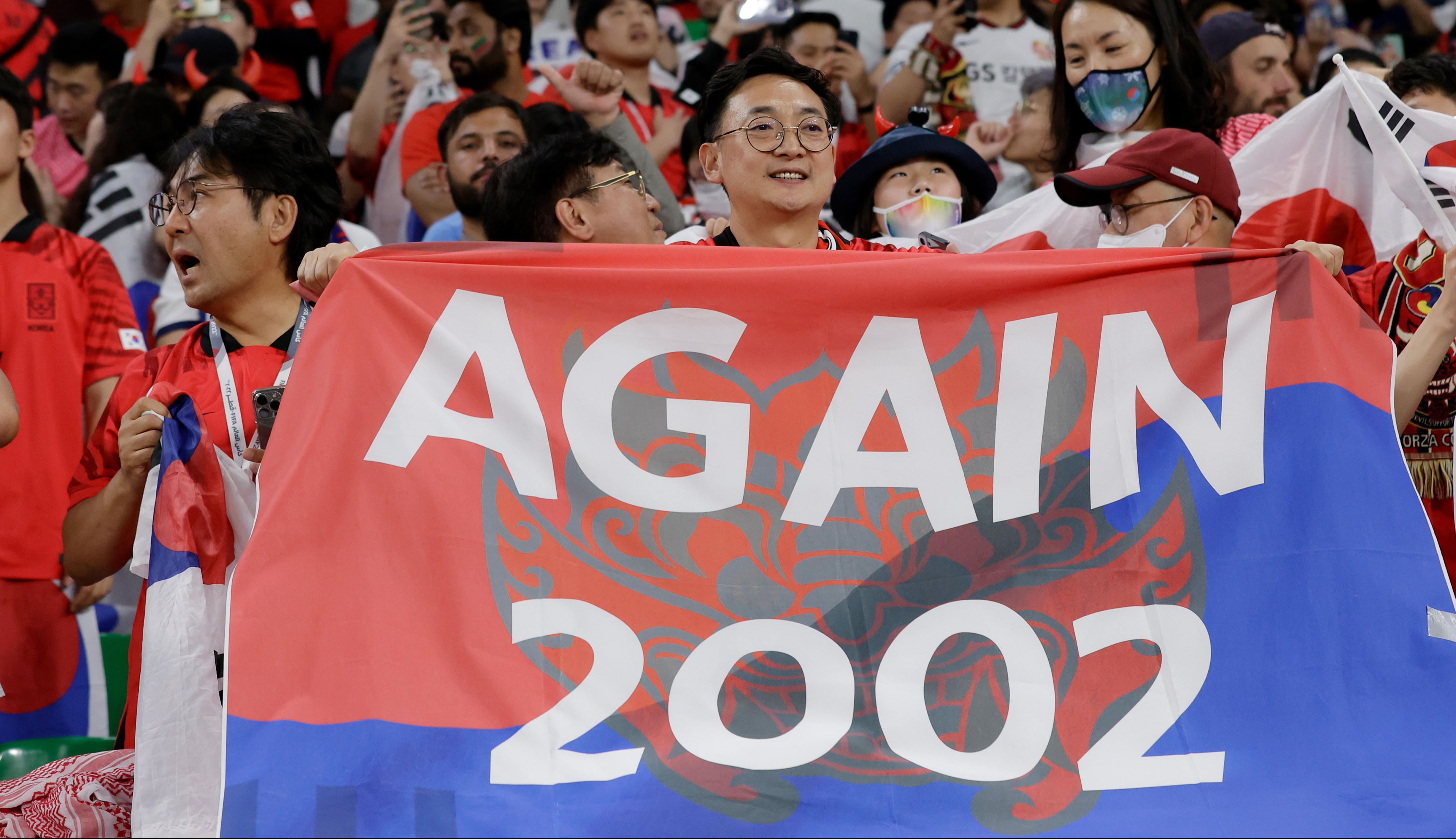 Excited sports fans hold a red and blue banner with the text 'AGAIN 2002'.